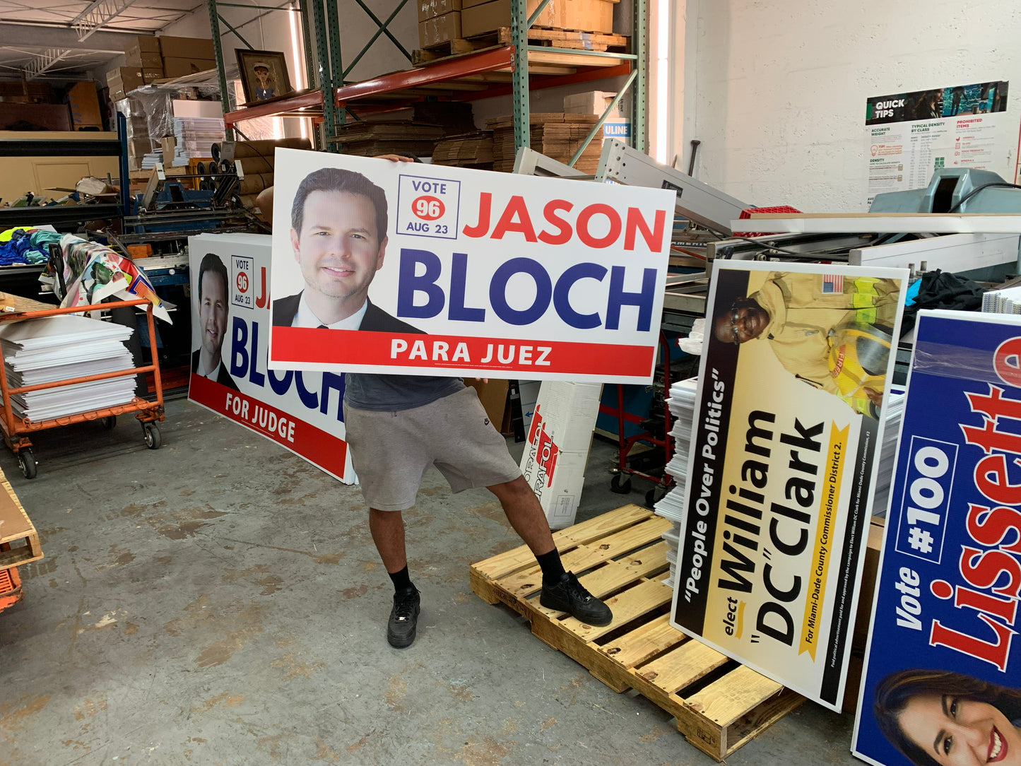 Person holding a political sign for Jason Bloch in a warehouse setting with various other signs.