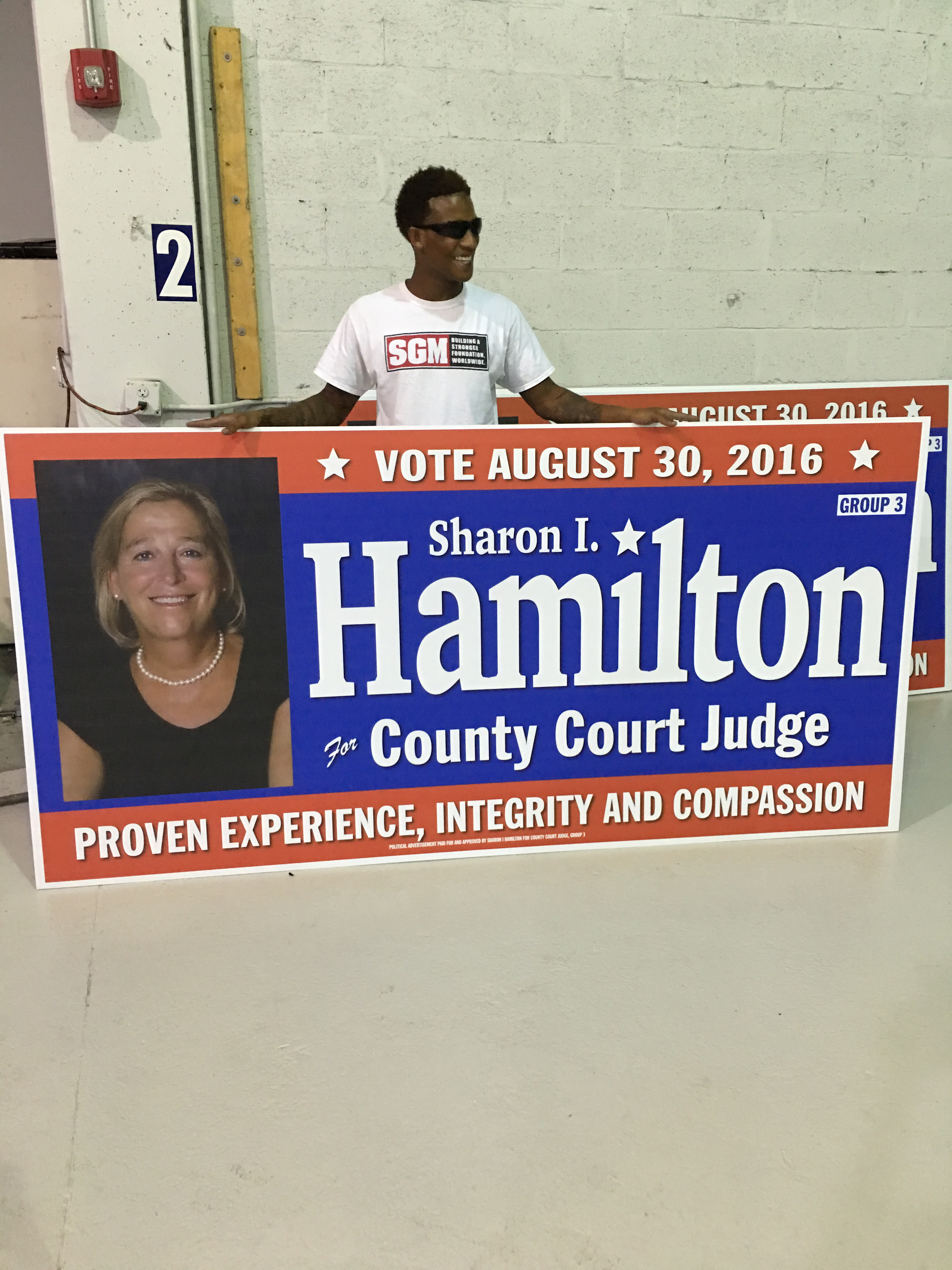 Person standing behind a political campaign sign for Sharon I. Hamilton for County Court Judge.