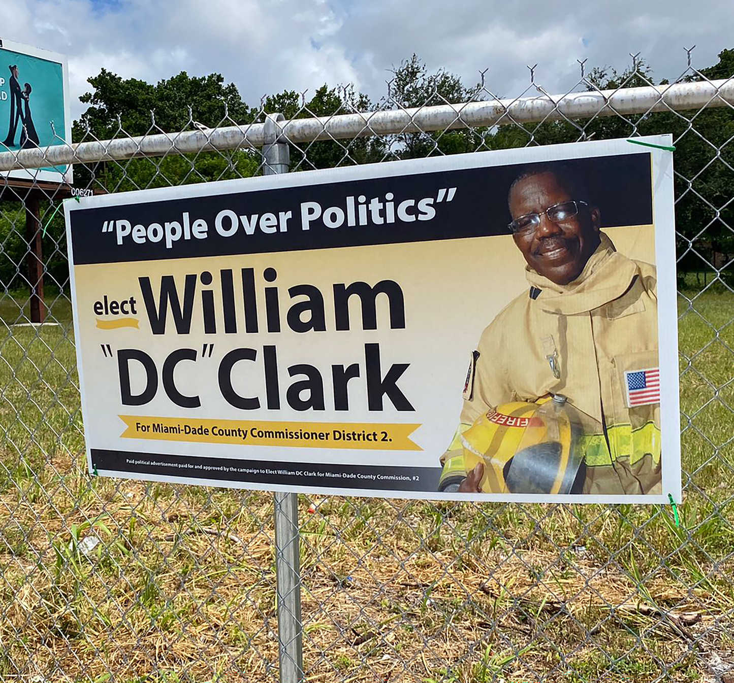 Campaign sign for William 'DC' Clark with a photo of a person in a uniform, placed against a chain-link fence.